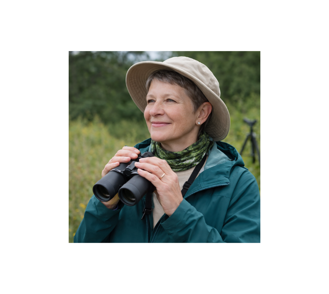Woman in outdoor setting with binoculars, wearing a head and neck buff with images of chickadees round her neck