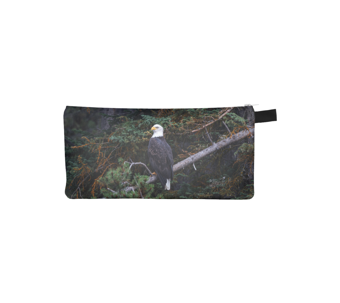 This image is of a photo of a bald eagle perched on a grey fallen tree branch. The background is of trees and branches in various tones of green, gold, purple and grey. The bald eagle has his back to us and its head is turned slightly, as he is looking towards you.