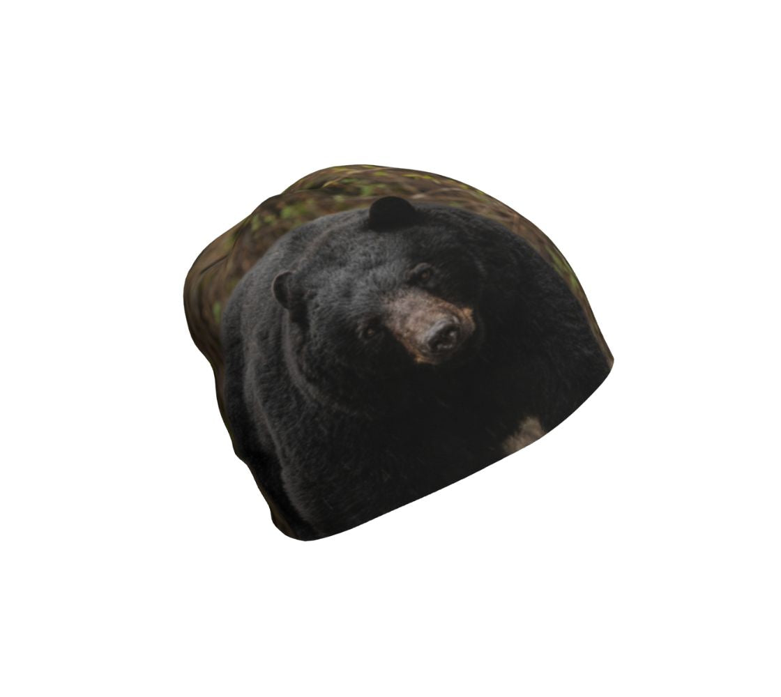 This is an image of a beanie featuring a photo of a big black bear head. The background is the mottled green forest behind him.