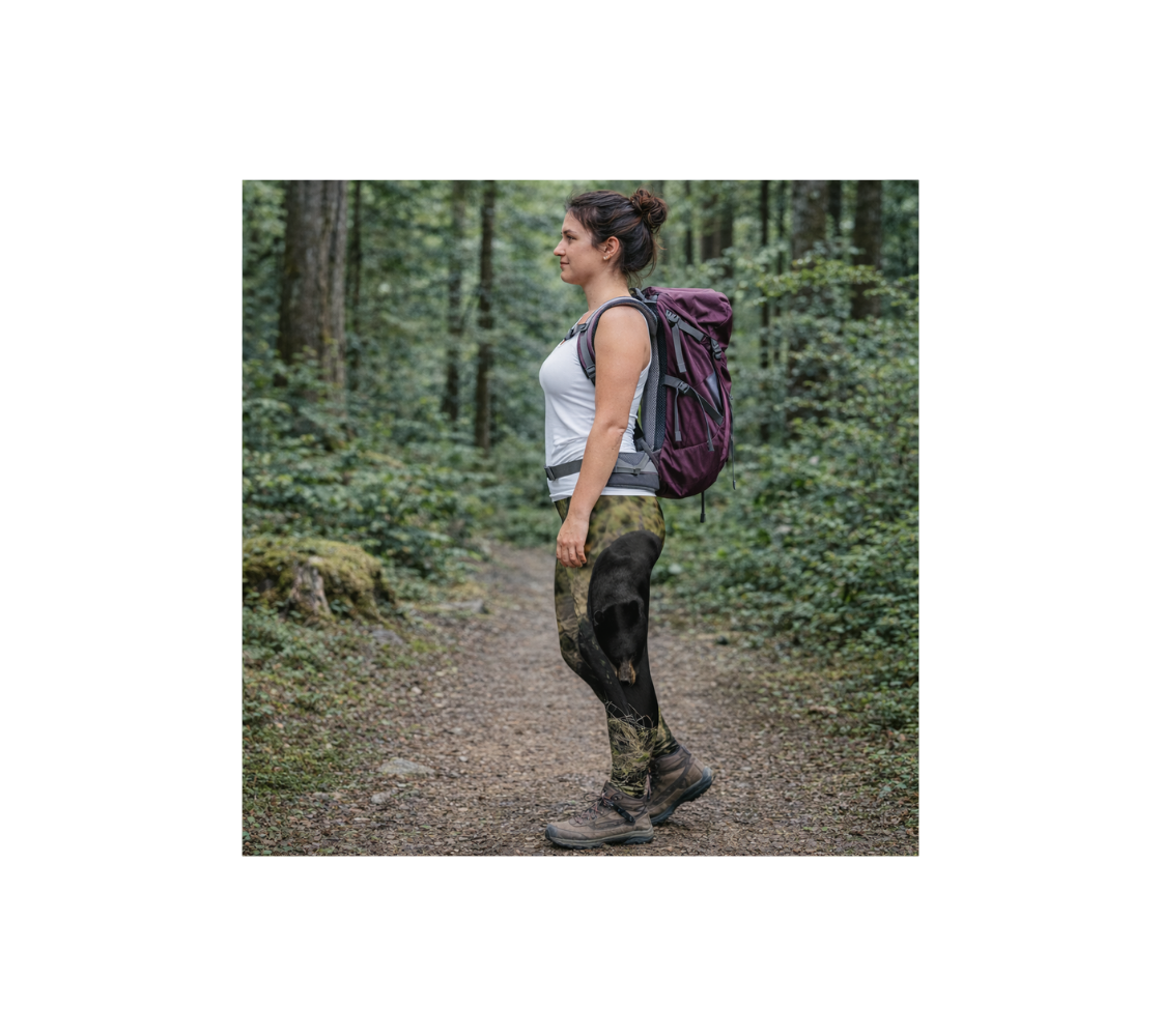 A Woman hiking in a forest wearing a pair of classic leggings with an image of a black bear design with a green background.