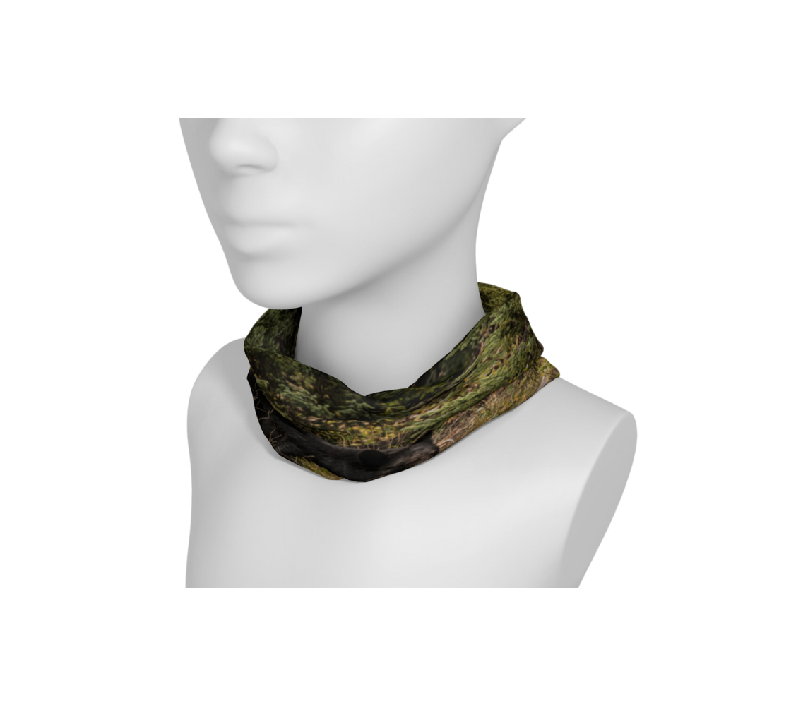 This is a photograph of a black bear on a head and neck band, worn as a neckband on a mannequin. The large black bear is walking along a hill covered in green grasses and bushes with a old grey log lying in front of him.