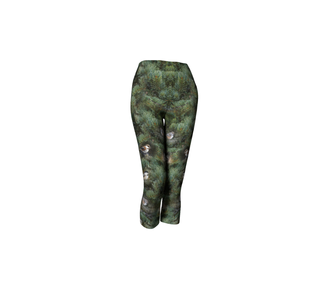 This is an image of a front profile of a pair of 3/4 length capris, featuring a photo with green tree boughs in the background with a repeating photo collage of the four different chickadees of Western Canada in their colorful black, brown, white, rust and grey tones.