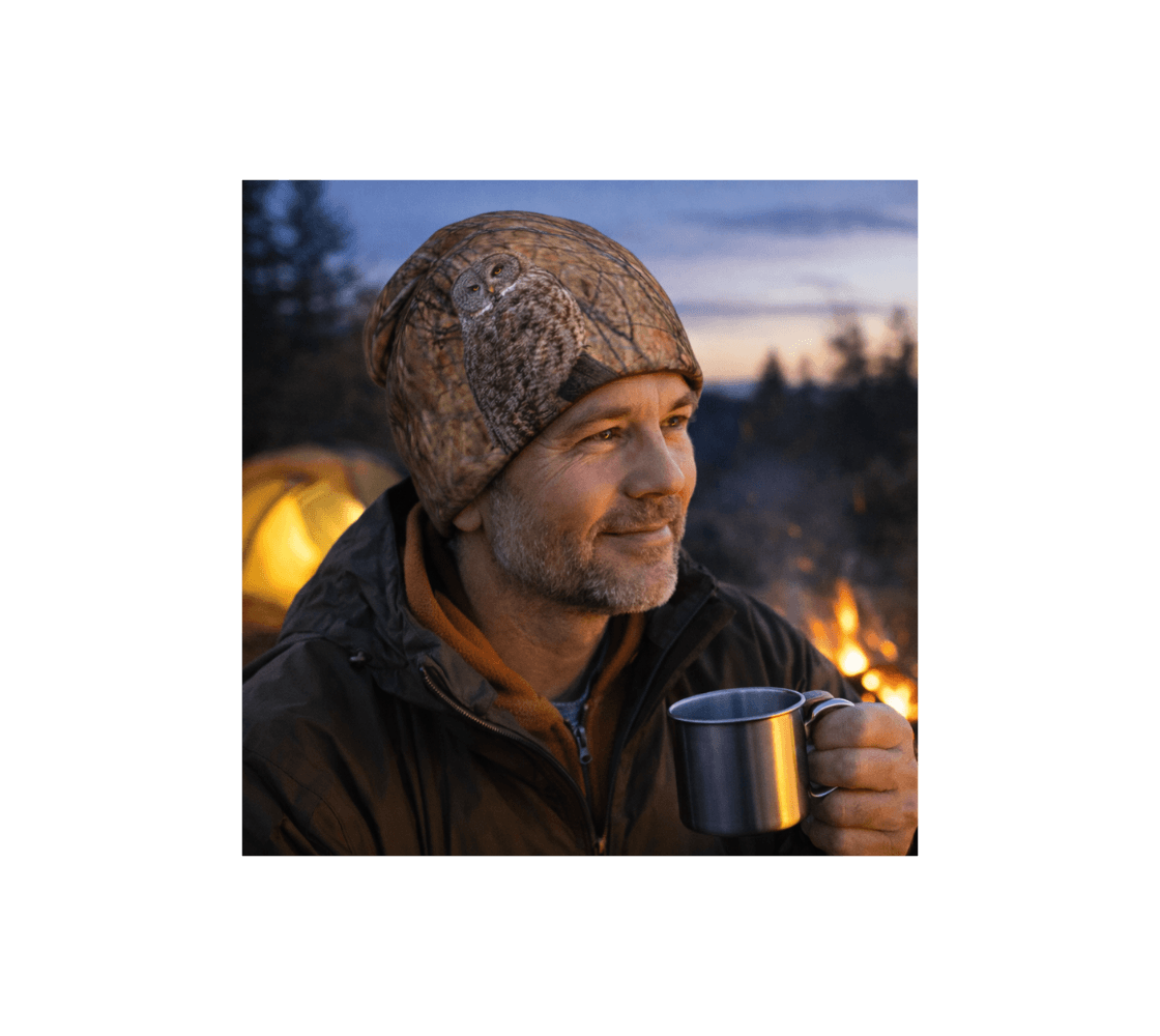 A man wearing a great gray owl bamboo-lined beanie from our wildlife photography holding a mug by a campfire.