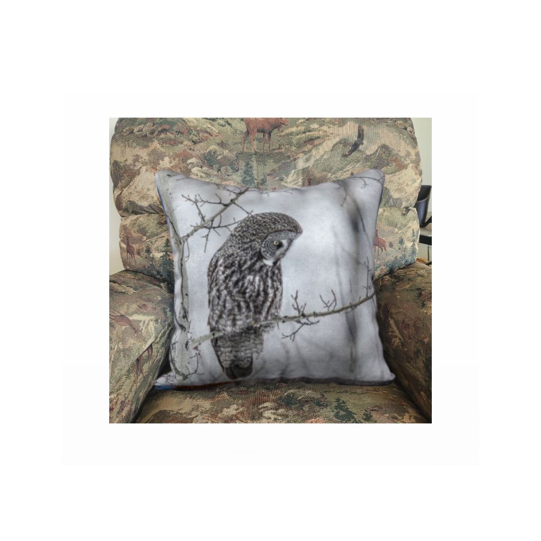 A 18 x 18 inch plush velveteen cushion cover features a photo of a great grey owl in the foggy morning. The background is white and grey, with blurred tree branches due to the fog and frost, with a great grey owl perched on branch. This image is of the cushion cover on a pillow form in a chair.