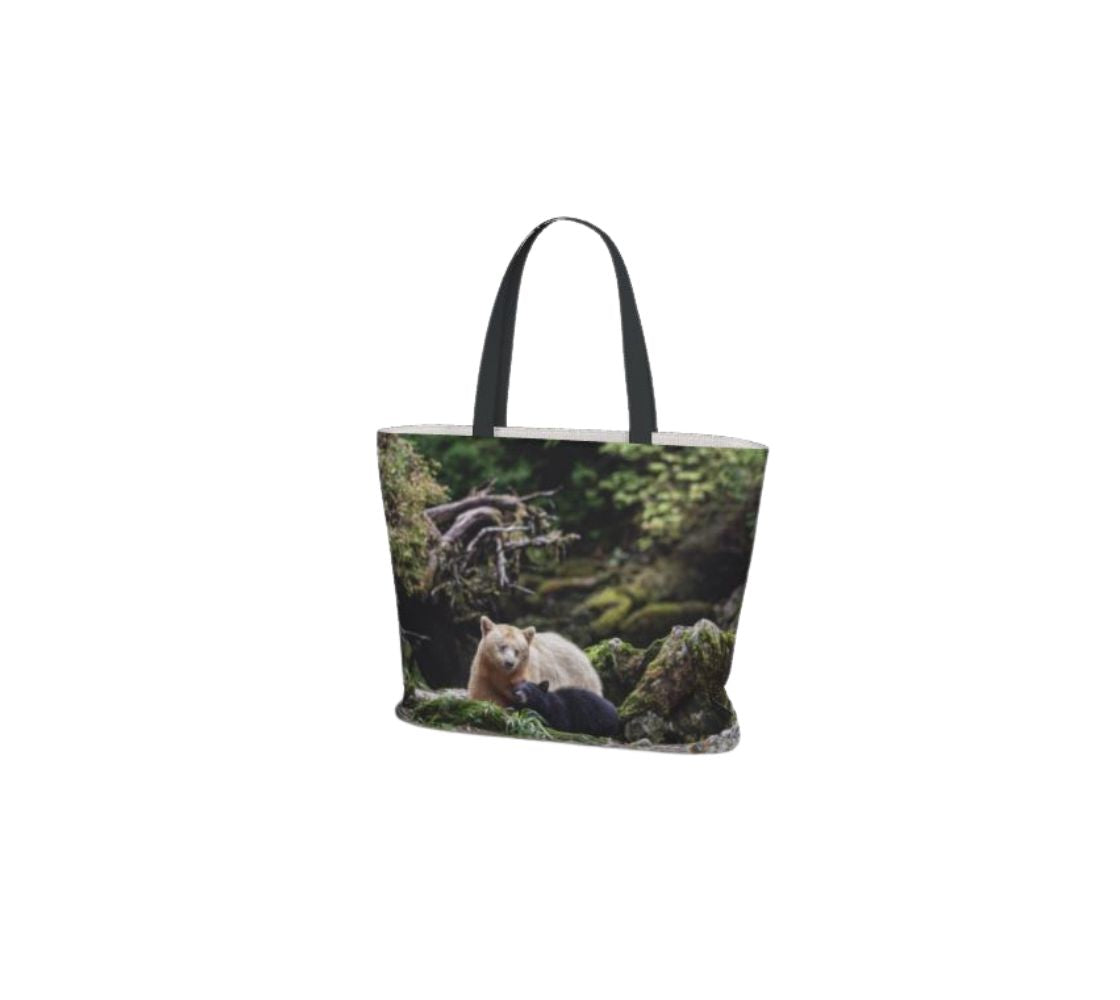 This large canvas tote bag has a photo of a white mama spirit bear with her black bear cub beside her. The background is grey rocks and tree roots covered in green moss. The bag has two 1.5" wide black straps with a white interior.