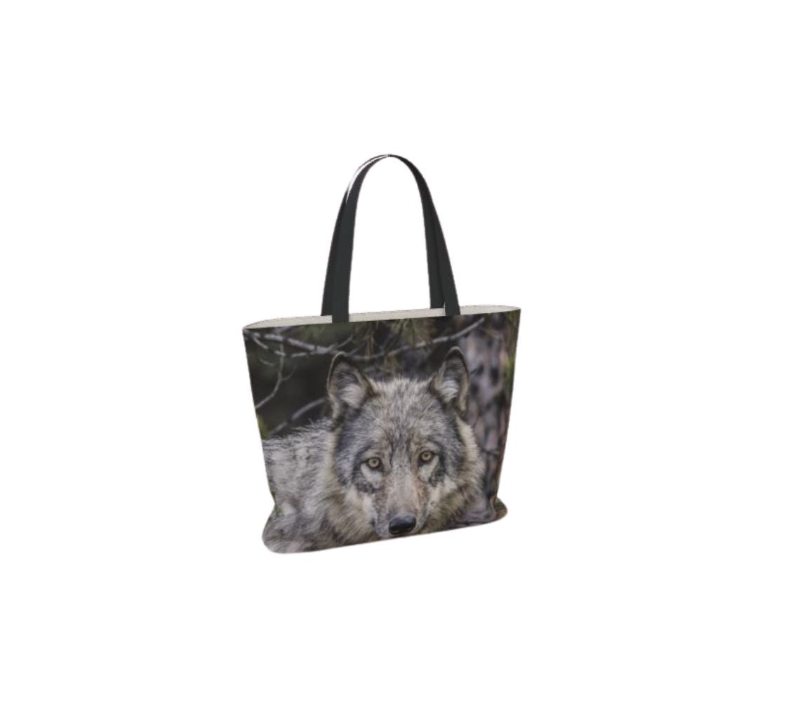 This image is of a large canvas tote bag featuring a photo of a grey wolf head shot. The background is of evergreen trees and grey bushes, while the wolf's coat is full and fluffy in various tones of grey, white and black with beautiful yellow eyes looking directly at you. The bag has two 1.5" wide black straps with a white interior.