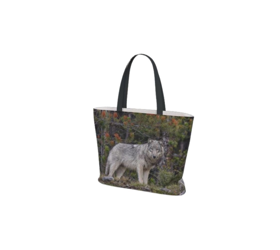 This image is of a large canvas tote bag featuring a photo of a grey wolf. The background is of evergreen trees and grey bushes, while the wolf's coat is full and fluffy in various tones of grey, white and black with beautiful yellow eyes looking directly at you. The bag has two 1.5" wide black straps with a white interior.