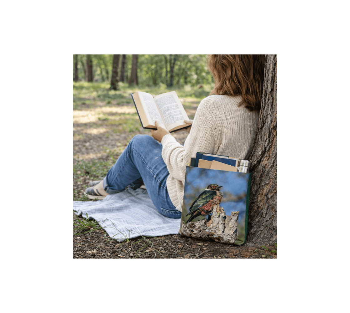 A woman sitting by a tree reading a book with a Lewis's woodpecker medium reusable tote bag from our real wildlife photography with books inside it.