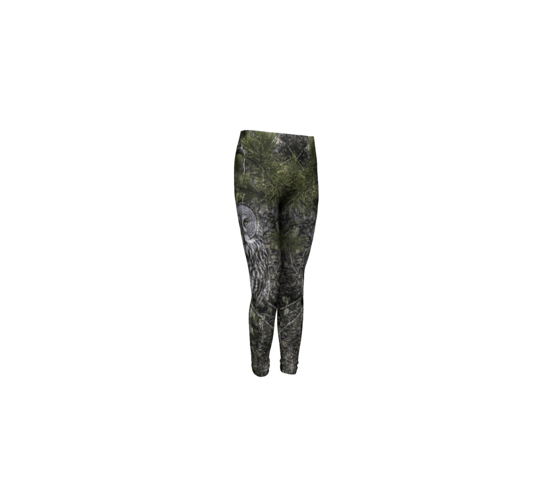 This is a front profile of a pair of kids leggings with a photo of an owl on the side of each leg. The background is green pine boughs and lichen hanging from the grey pine tree branches, with a great grey owl with black, grey and white feathers, and yellow eyes, perched on a branch.