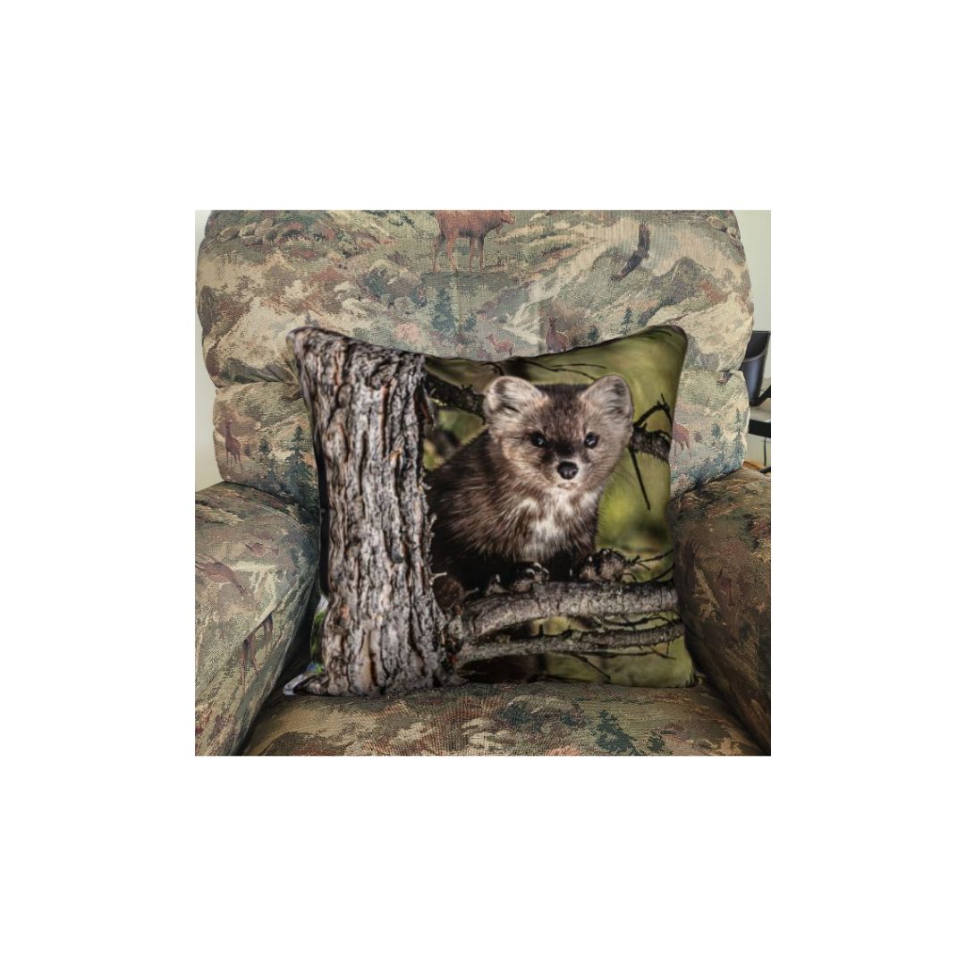 This 18 x 18 inch plush velveteen cushion cover features a photo of a dark brown pine marten on a light brown tree branch looking at you. The cushion cover background is mottled green and black. This image is of the cushion cover on a pillow form in a chair.