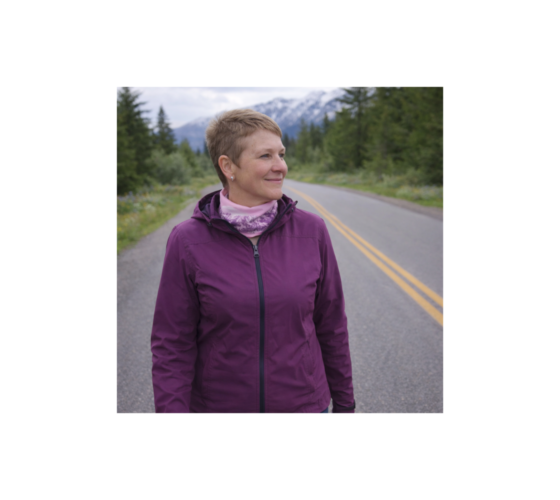Woman standing on the side of a road with mountains background wearing a head and neck buff around her neck with our photograph of a pink mountain sunset design.