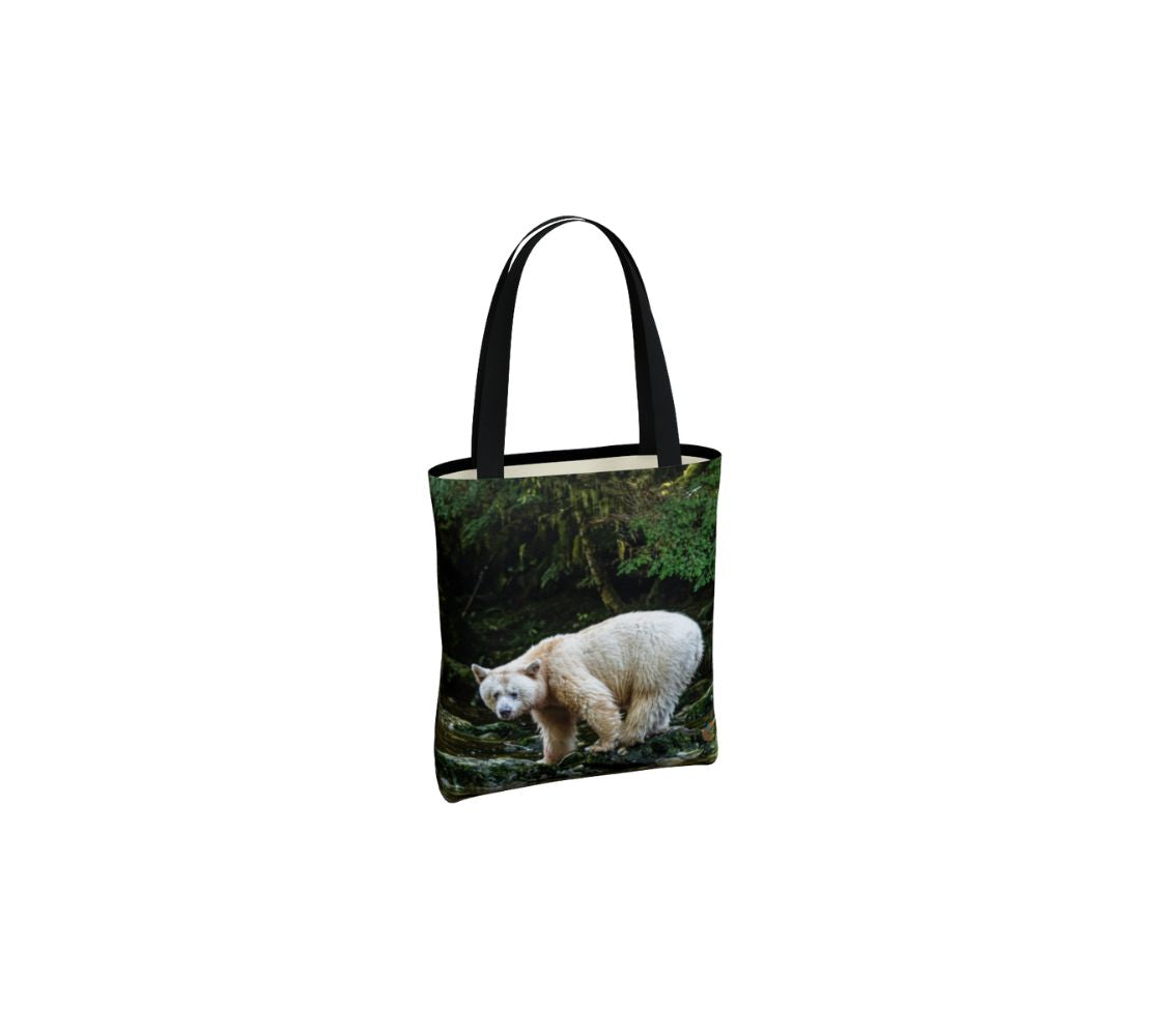 This side of the canvas tote bag has a white spirit bear standing on a moss covered rock in a flowing stream. Background is green forest trees with dark shaded areas. The bag has two 1.5" wide black straps with a white interior.