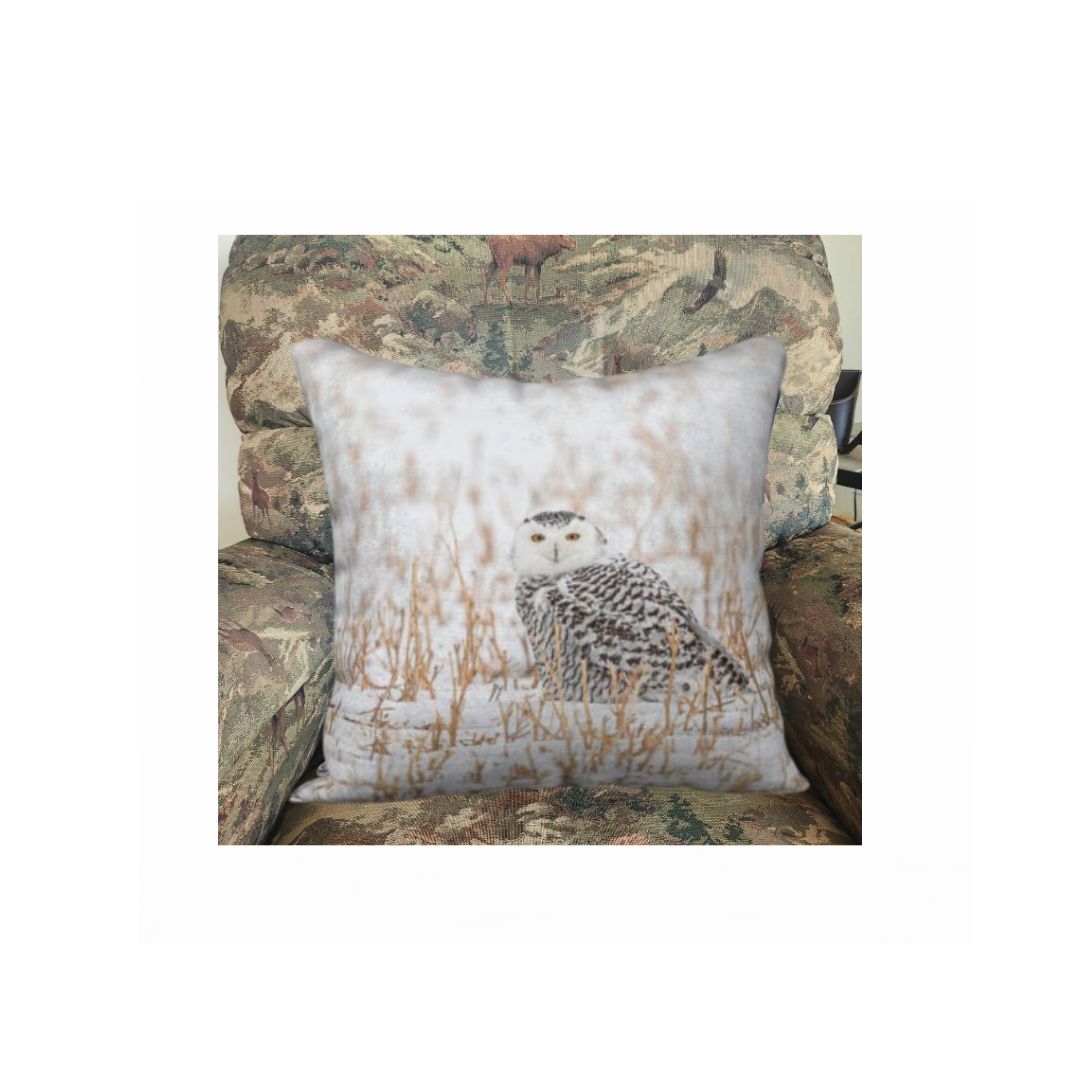 A 18 x 18 inch plush velveteen cushion cover features a photo of a snowy owl. The image is of a snowy owl looking at you. It is on the ground in a white snow covered field surrounded by gold colored field stubble sticking up out of the ground.