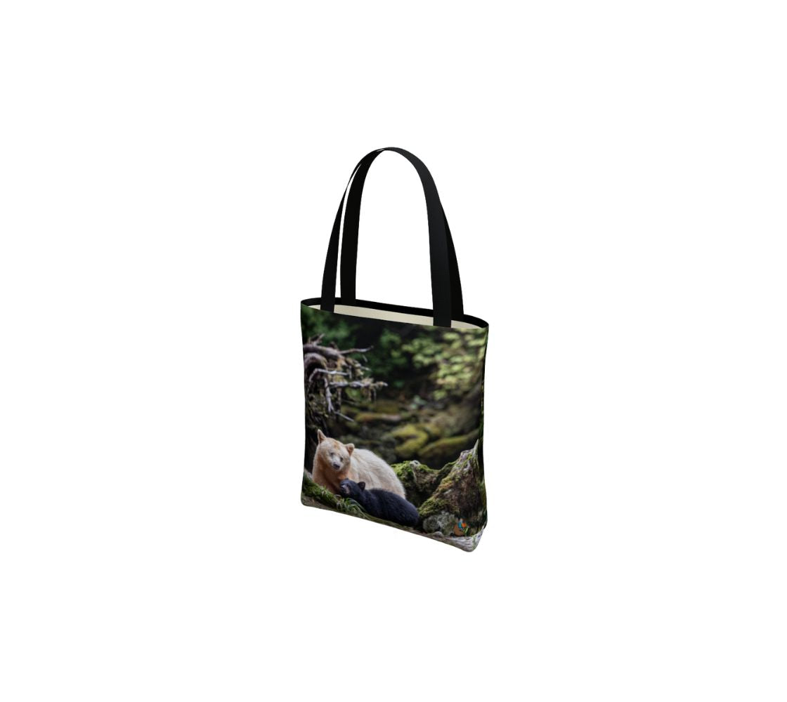 This canvas tote bag has a photo of a white mama spirit bear with her black bear cub beside her. The background is grey rocks and tree roots covered in green moss. The bag has two 1.5" wide black straps with a white interior.