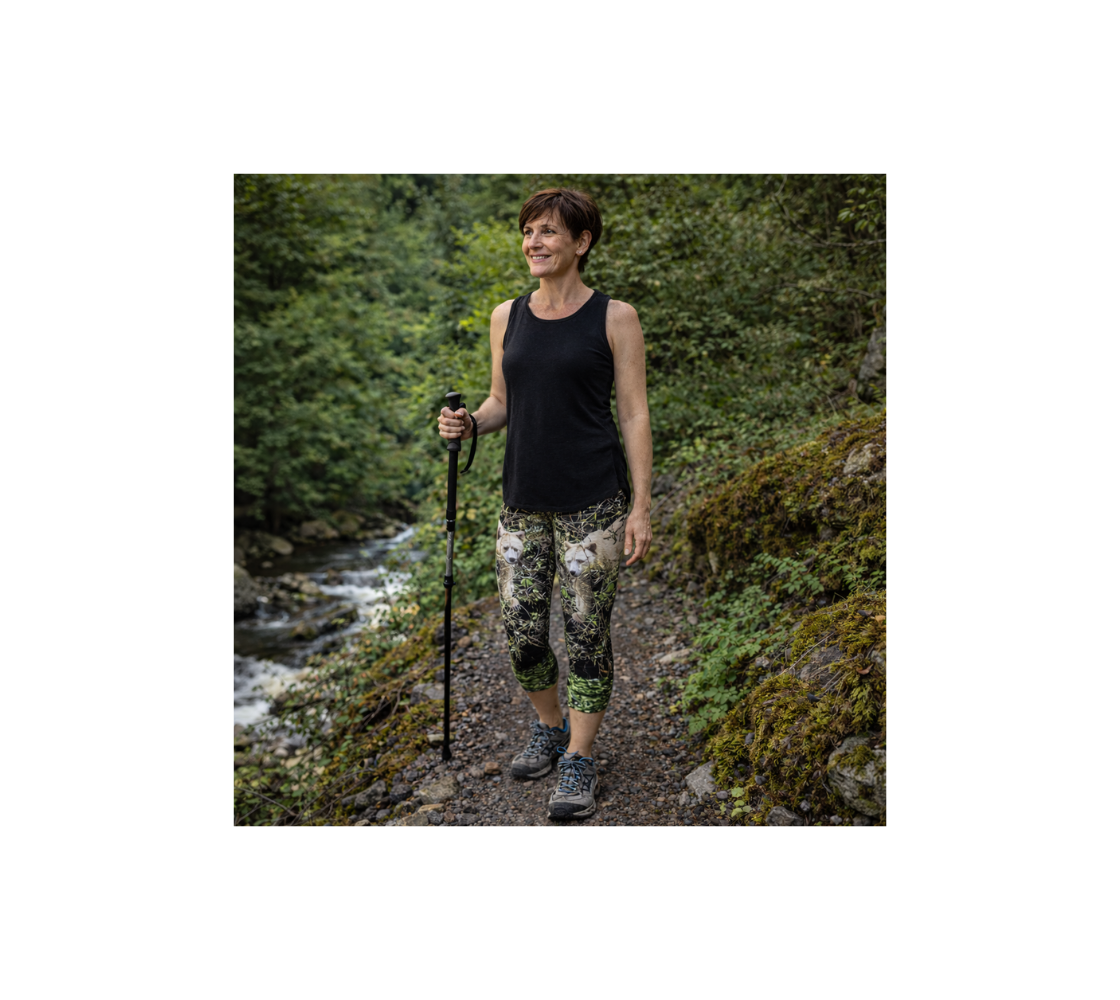 A Woman hiking in a forest wearing a pair of classic capri leggings with an image of a spirit bear design with a green background standing by a stream.