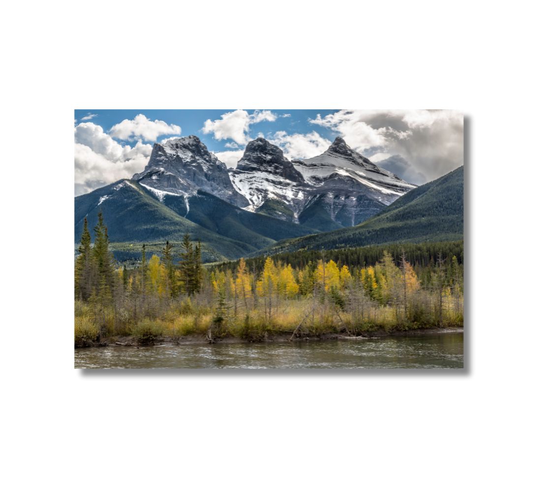 This is a 16 x 24 landscape canvas print. This is a photo of The Three Sisters mountain at Canmore, Alberta, photographed in the autumn. The background is of green trees on the mountain, blue sky with white clouds and yellow trees in the foreground with the river running along the front of the scene.