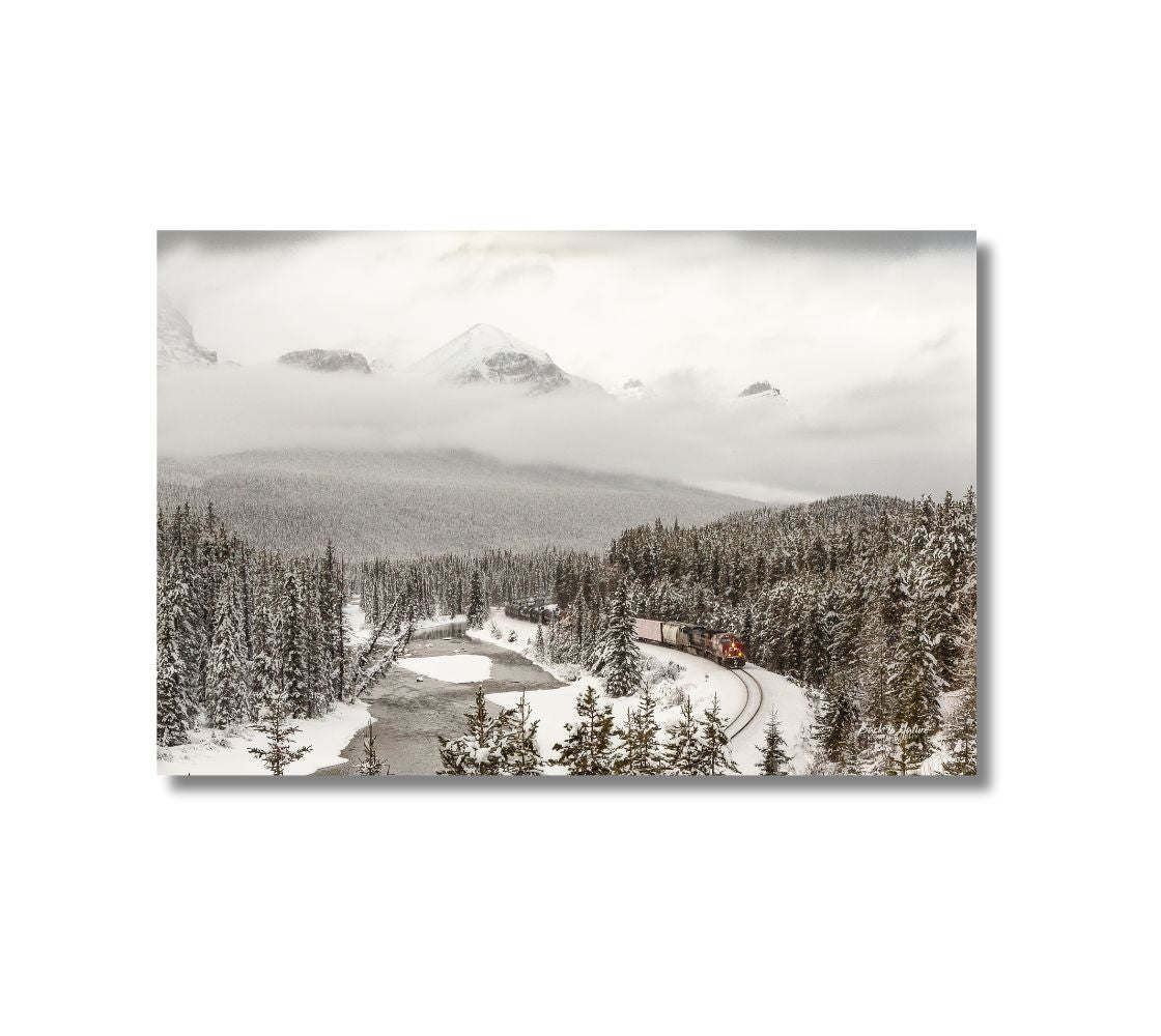 This is a 16 x 24 landscape canvas print. This is a photo of a train with a red engine and other cars behind coming around Morant's  Curve in Banff National Park. It is a cloudy winter scene and the background is cloud covered mountains, evergreen trees blanketed in snow, and the open, but snow covered river next to the railway track.