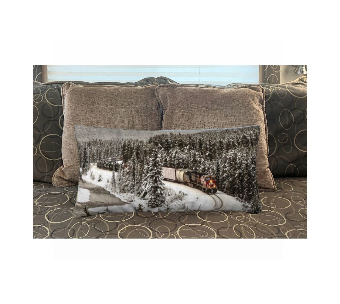 This 12x24 inch plush velveteen cushion cover features a photo of a trian, with a red engine coming around Morant's Curve along a flowing river in the mountains during the winter. The background is grey, with dark snow-topped evergreen trees. This image is of the cushion cover on a pillow form on a bed.