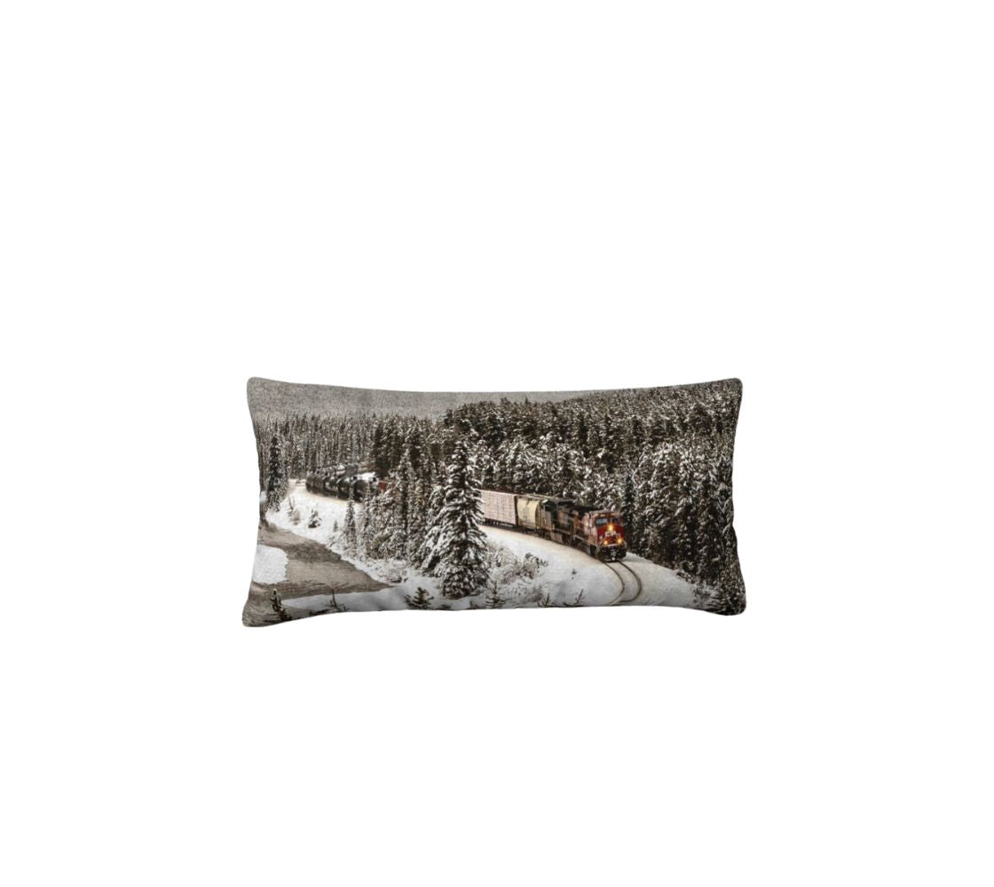 This 12x24 inch plush velveteen cushion cover features a photo of a trian, with a red engine coming around Morant's Curve along a flowing river in the mountains during the winter. The background is grey, with dark snow-topped evergreen trees.