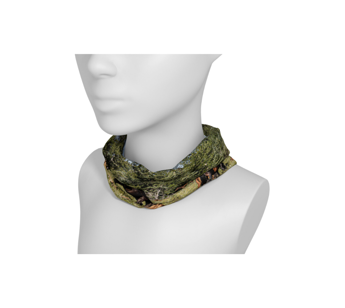 This is a head and neck band of a herd of wild horses on a mannequin head worn as a scrunched up neckband, featuring a photograph of wild foals lying in wildflowers in the summer, with a herd of wild horses grazing in a field surrounded by trees with a blue sky. Color tones include greens, browns, blue and black.