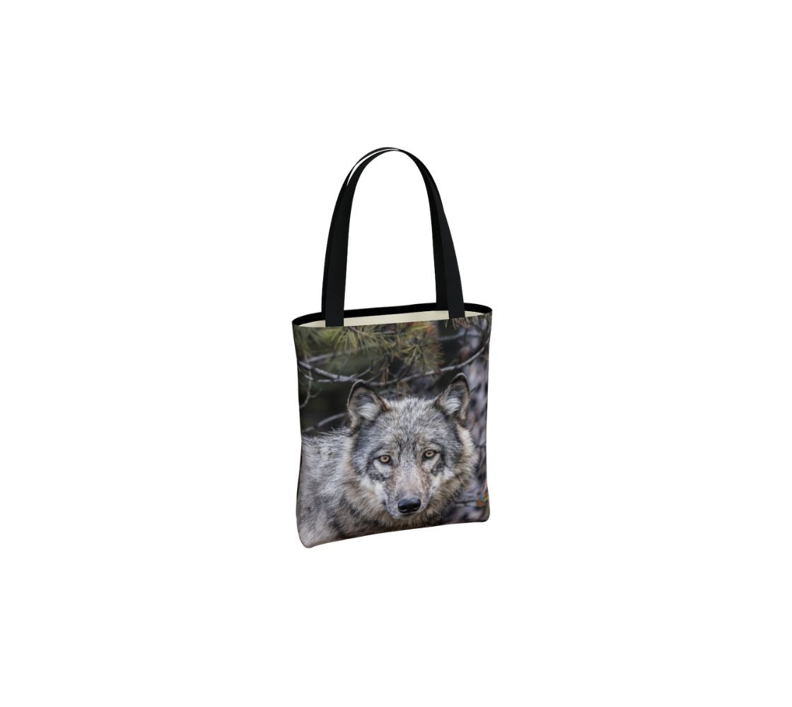 This image is of a small canvas tote bag featuring a photo of a grey wolf head shot. The background is of evergreen trees and grey bushes, while the wolf's coat is full and fluffy in various tones of grey, white and black with beautiufl yellow eyes looking directly at you. The bag has two 1.5" wide black straps with a white interior.