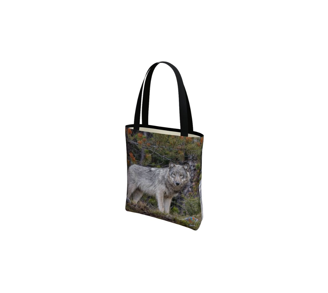 This image is of a small canvas tote bag featuring a photo of a grey wolf. The background is of evergreen trees and grey bushes, while the wolf's coat is full and fluffy in various tones of grey, white and black with beautiufl yellow eyes looking directly at you. The bag has two 1.5" wide black straps with a white interior.