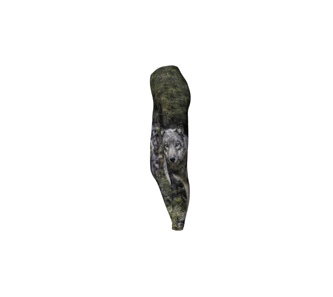 This is a side profile of a pair of yoga leggings with a photo of a wolf ion the side of each leg, with a 4.5 inch wide waistband. The background is of evergreen trees and grey bushes, while the wolf head is full and fluffy in various tones of grey and white with beautiufl yellow eyes looking directly at you.
