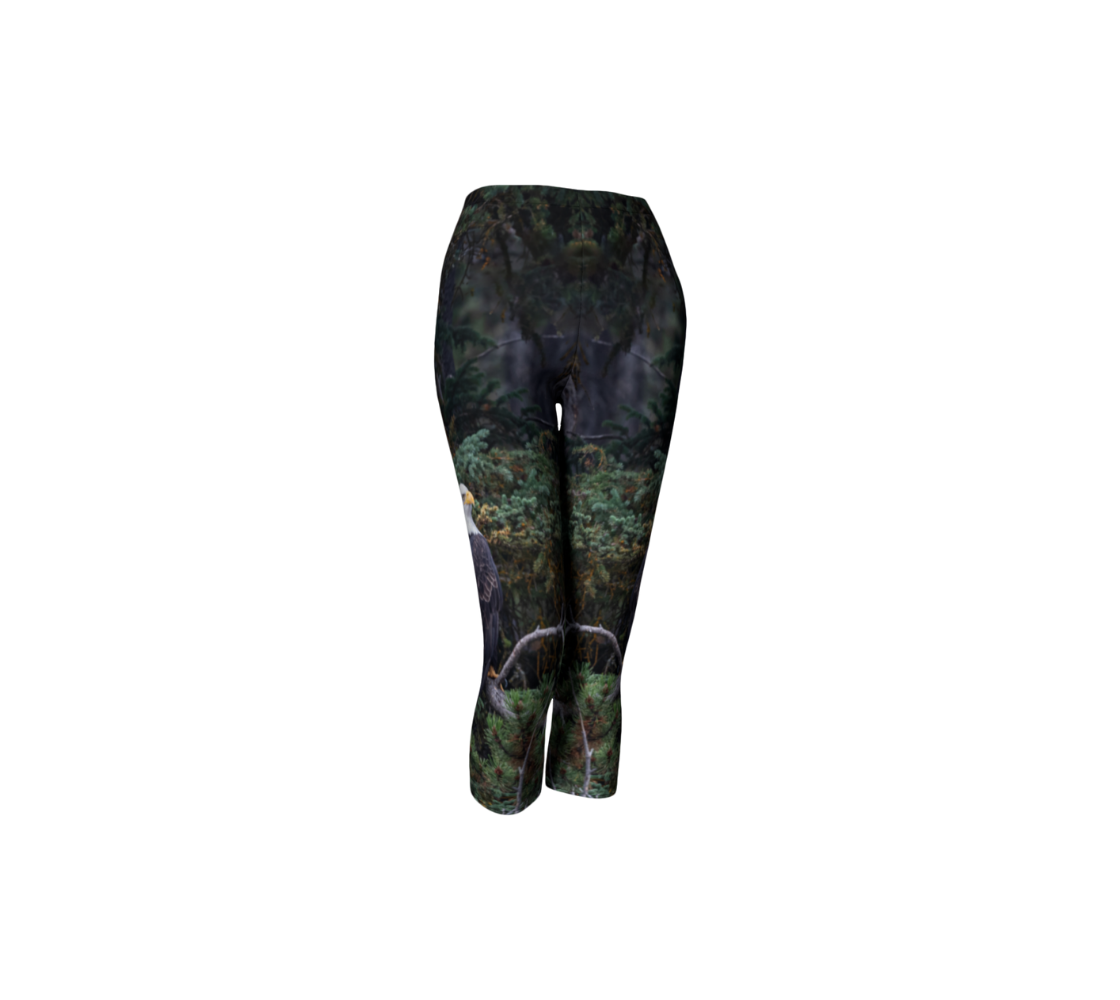 This is a front profile of a photo of a bald eagle on each side of a pair of 3/4 length capri leggings. The background is of trees and branches in various tones of green, gold, and grey. The bald eagle is perched on a fallen grey tree trunk with his back to us and its head is turned slightly, as he is looking towards you.
