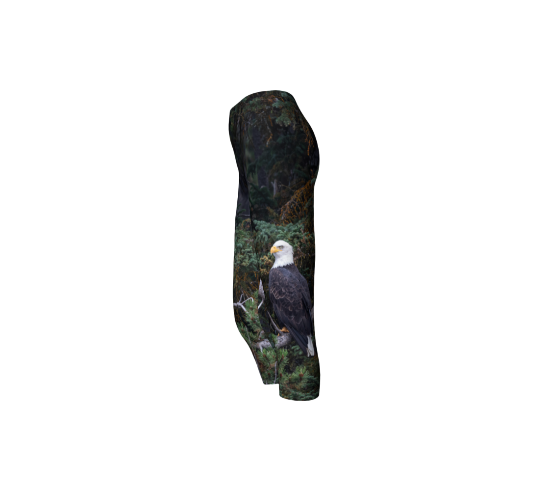 This is a side profile of a photo of a bald eagle on a pair of 3/4 length cpari leggings. The background is of trees and branches in various tones of green, gold, and grey. The bald eagle is perched on a fallen grey tree trunk with his back to us and its head is turned slightly, as he is looking towards you.
