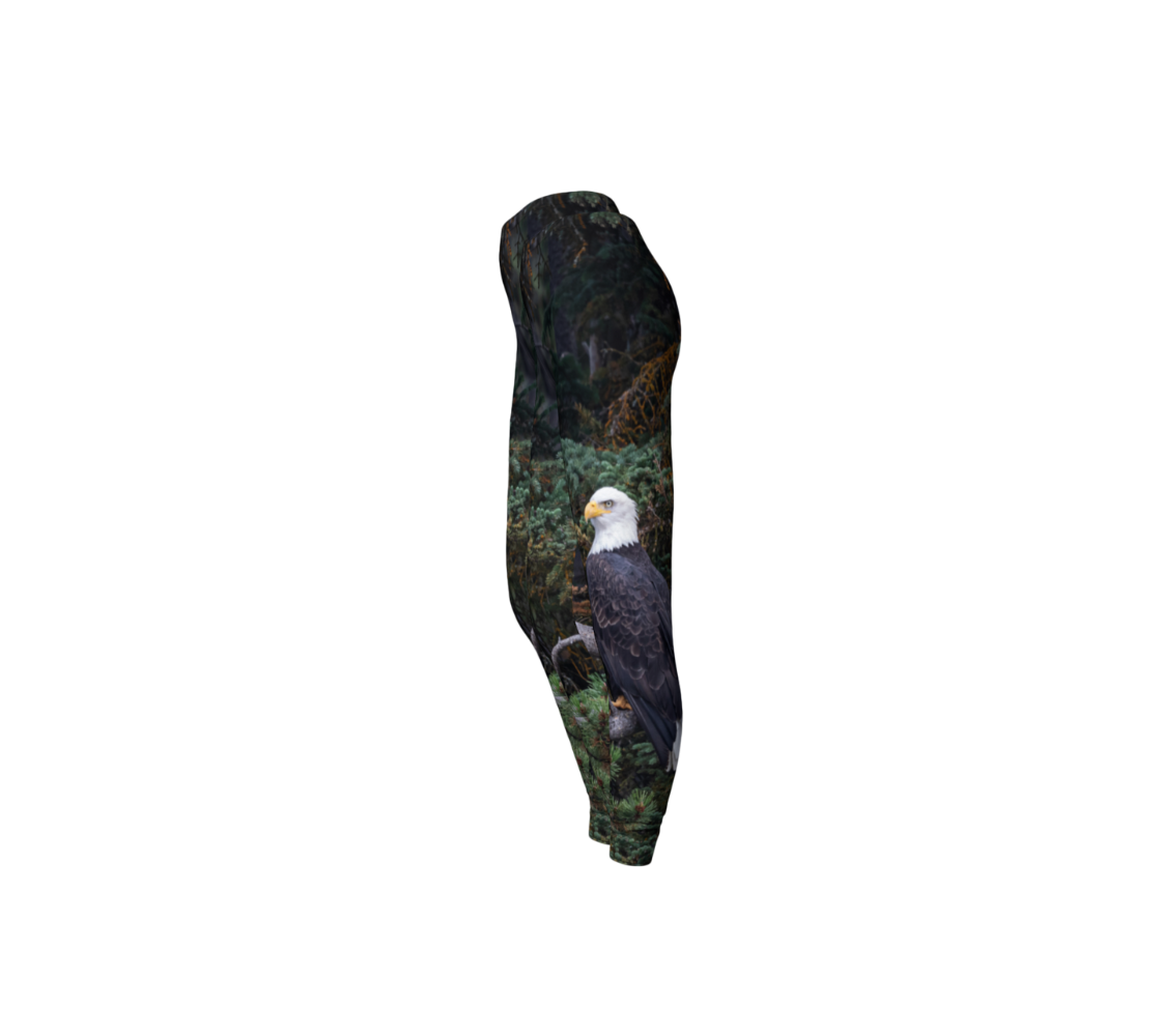 This is a side profile of a pair of classic leggings featuring a photo of a bald eagle on each leg, with a 1.5 inch wide elastic waistband. The background is of trees and branches in various tones of green, gold, purple and grey. The bald eagle is perched on a fallen grey tree trunk with his back to us and its head is turned slightly, as he is looking towards you.