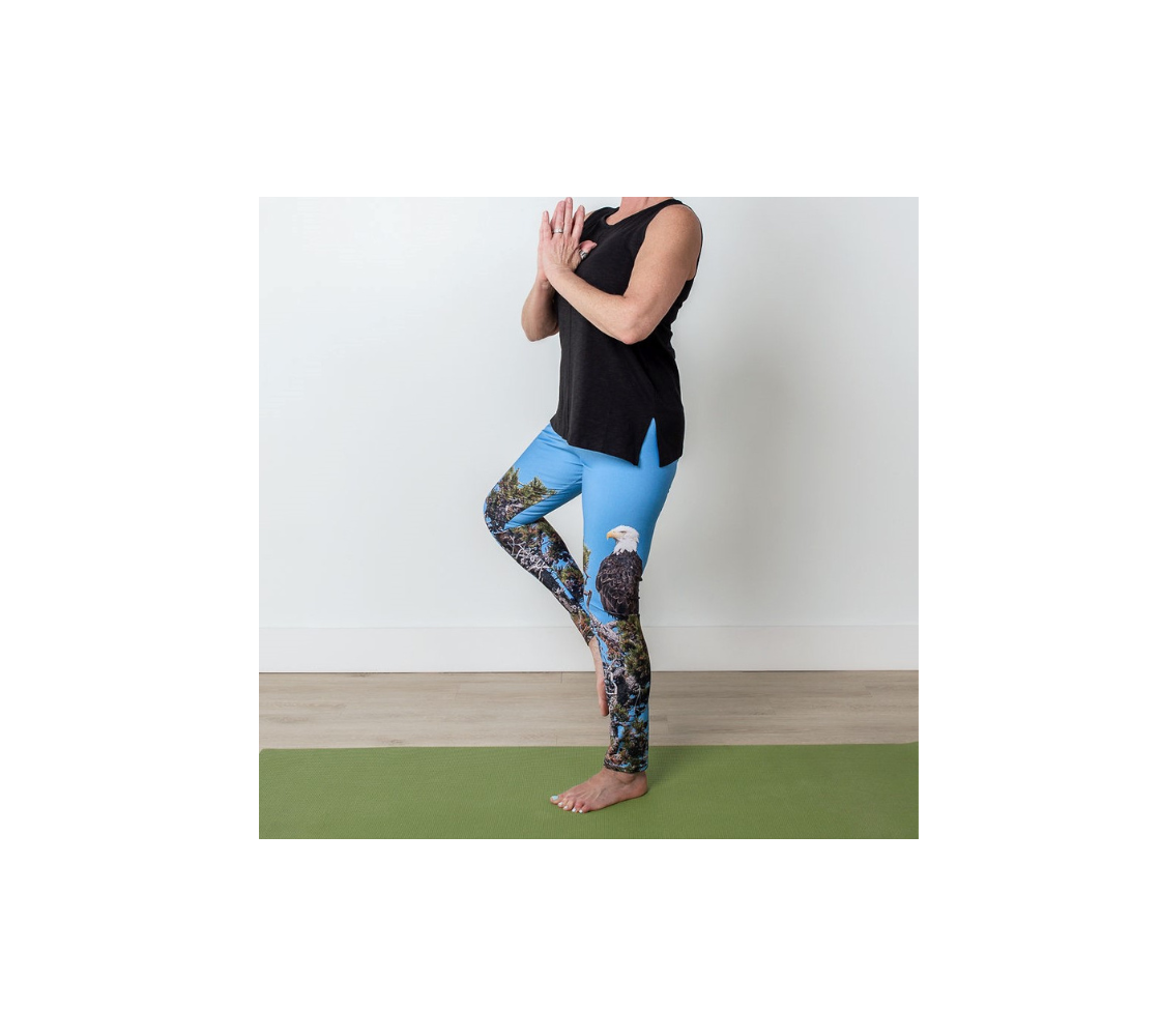 This is a side profile of a lady doing the tree yoga pose in a pair of yoga leggings featuring a photo of a bald eagle on the side of each leg, with a 4.5 inch high waist band. The background is a blue sky with a bald eagle perched on top of a pine tree.