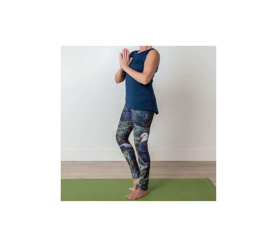 This is a side profile of a lady doing a yoga pose in a pair of yoga leggings featuring a photo of a bald eagle on each leg. The background is of trees and branches in various tones of green, gold, purple and grey. The bald eagle is perched on a fallen grey tree trunk with his back to us and its head is turned slightly, as he is looking towards you.
