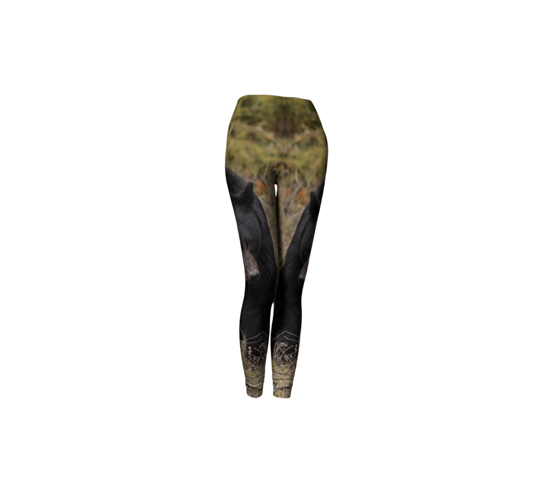 This is a front profile of a pair of classic leggings featuring a photo of a black bear on each leg, with a 1.5 inch wide elastic waistband. The mottled background is tall brown amd green grass. The large black bear is walking towards you.