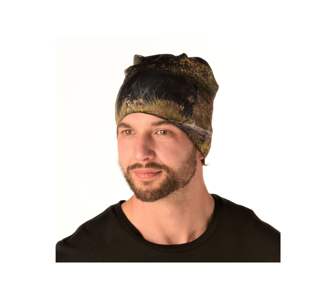 This is a photograph of a man wearing a photo of a black bear on a head and neck band, worn as a kerchief. The large black bear is walking along a hill covered in green grasses and bushes with a old grey log lying in front of him.