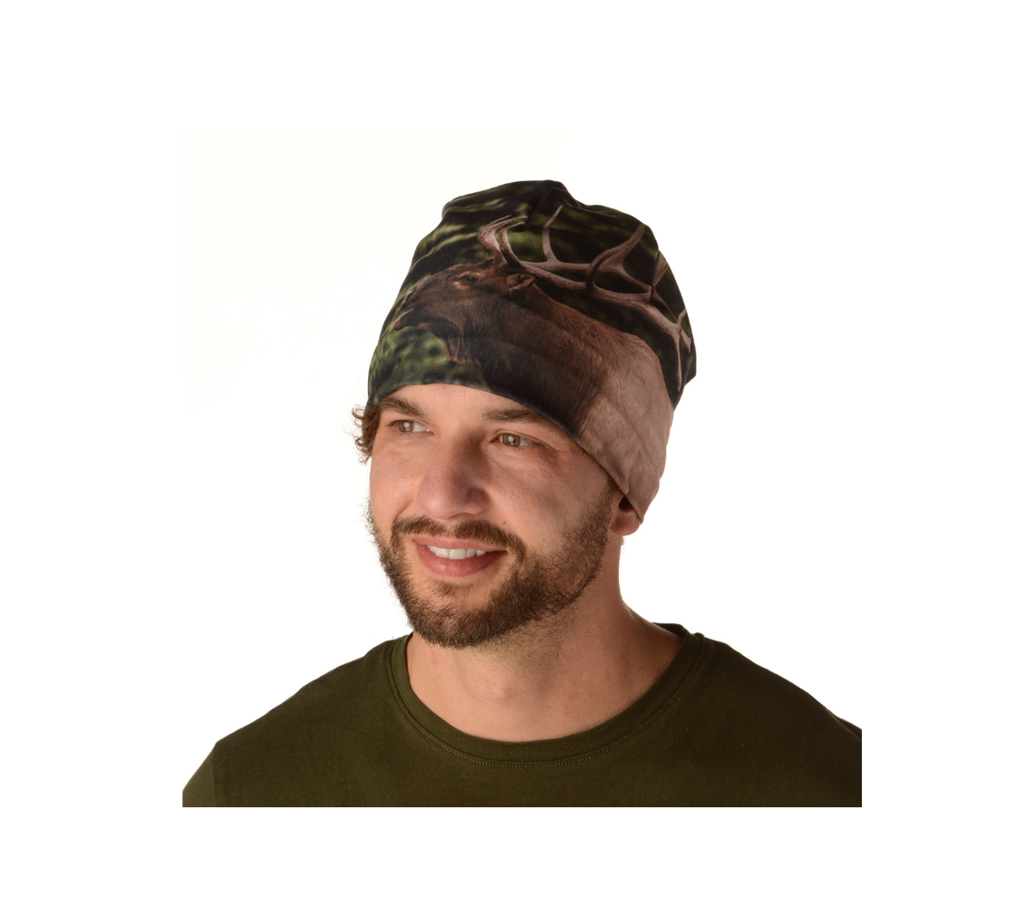This is a front portrait of a man wearing a photo of an elk on a beanie. The background is blurred evergreen trees with yellow dead grass. The dark brown and tan colored elk with its large antlers is raising its head as it bugles out its mating call.