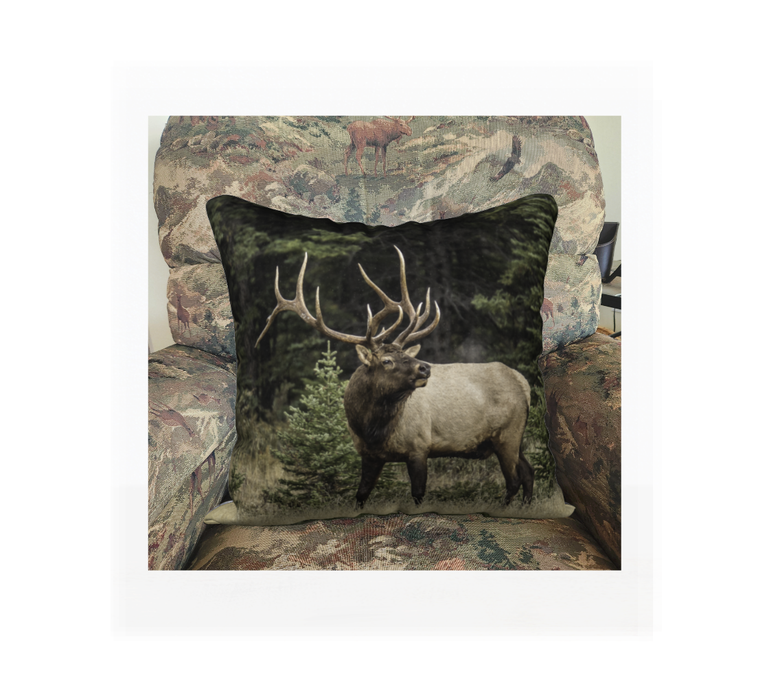 This 18 x 18 inch plush velveteen cushion cover features a photo of a two toned brown bull elk, with large antlers, standing with its head turned as it is bugling with exhaust coming from his mouth. The background is the green and golden tones of the autumn forest. This image is of the cushion cover on a pillow form in a chair.