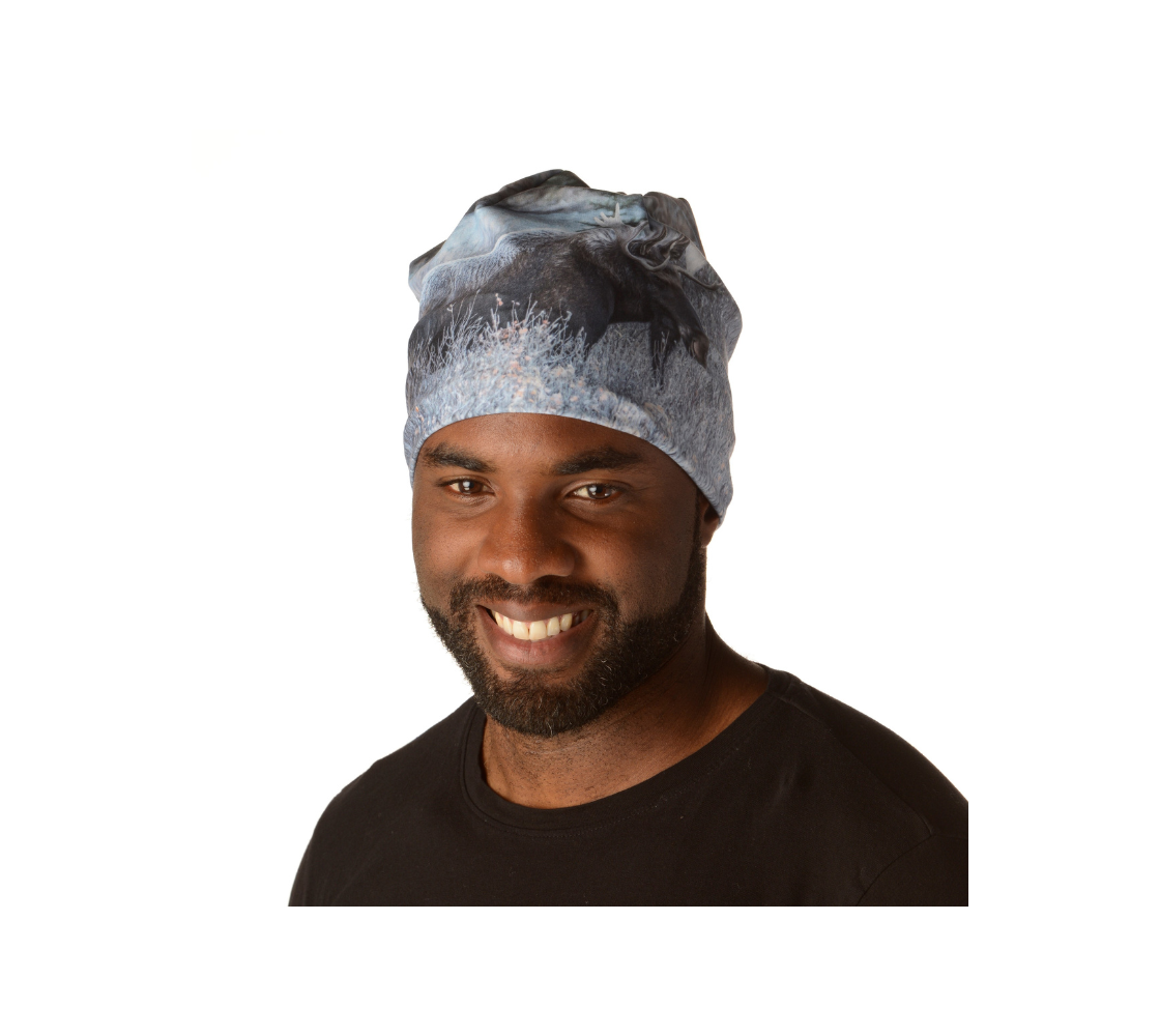 This is a front portrait of a man wearing a photo of a bull moose on a beanie. This image on the beanie shows a big dark brown bull moose standing in the frosty grey willows surrounded by evergreen trees. He is covered with a blanket of white frost on his back, shoulders and head.