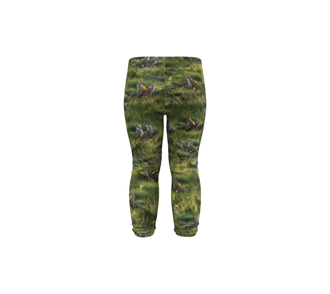 This is a rear profile of a pair of infant leggings featuring a collage of photos of red fox kits. The background is green grass, with various images of the brown and white fox kits scattered throughout.