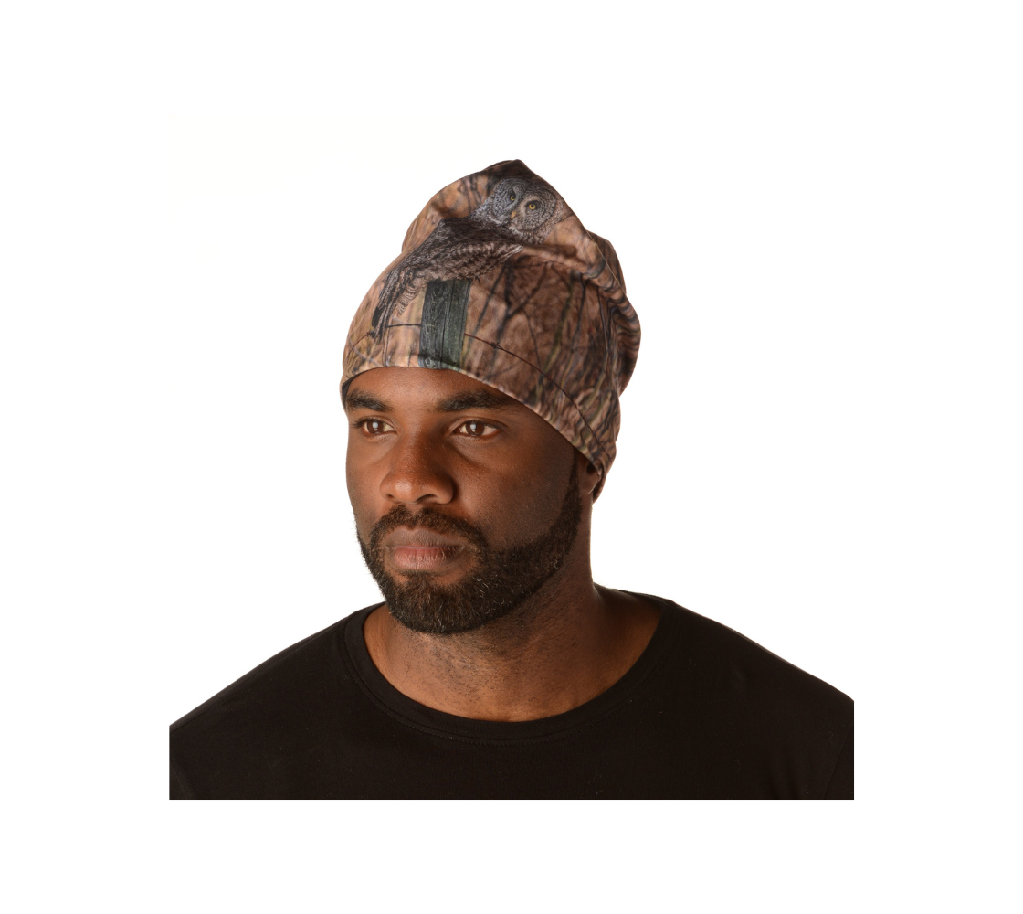 This is a front portrait of a man wearing a beanie with a photograph of an owl on it. The background is blurred brown twigs and tall yellow dead grasses. The brown and tanned colored owl is sitting on an old grey fence post looking at you.