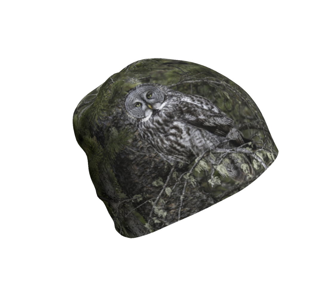 This is a photograph of a great grey owl on a beanie. The background is green pine boughs and lichen hanging from the grey pine tree branches, with a great grey owl with black, grey and white feathers, and yellow eyes, perched on a branch.