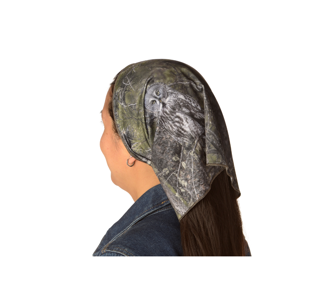 This is an image of a lady wearing a photo of a great grey owl on a head and neck band, worn as a kerchief. The background is green pine boughs and lichen hanging from the grey pine tree branches, with a great grey owl with black, grey and white feathers, and yellow eyes, perched on a branch.