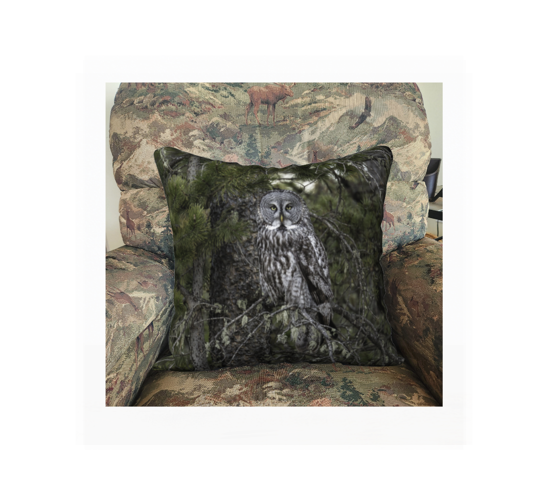This 18 x 18 inch plush velveteen cushion cover features a photo of a great grey owl . The background is green pine boughs and lichen hanging from the grey pine tree branches, with a great grey owl with black, grey and white feathers, and yellow eyes, perched on a branch. This image is of a cushion cover on a pillow form in a chair.