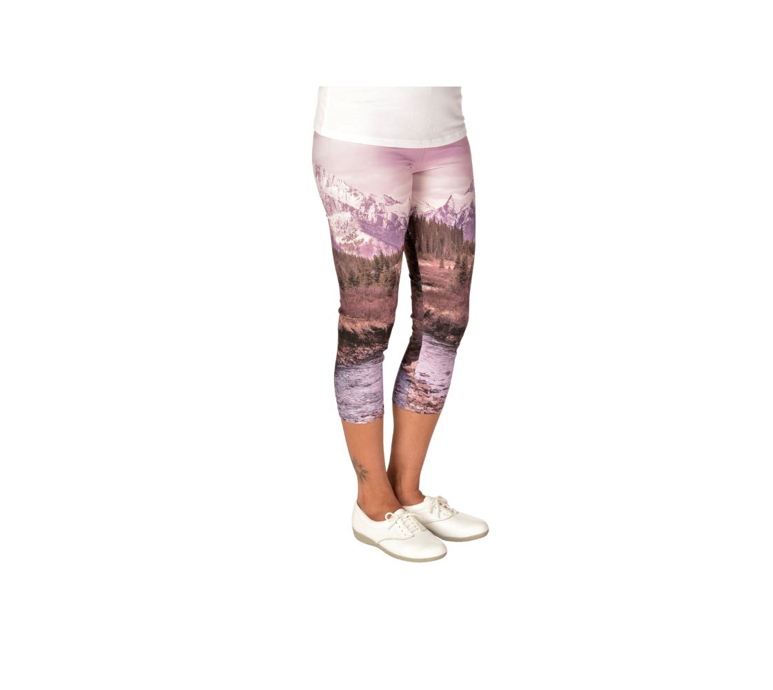 This is a side profile of a lady wearing a pair of 3/4 length capri leggings featuring a photo of a pink mountain scene. The image is of a snow covered mountain in the far distance with a meandering stream winding its way through the fall colored willow and forest of evergreen trees towards the mountain. A special filter used when taking this photograph created an over all pink hue to the image that really accentuated the sunset light at the time.