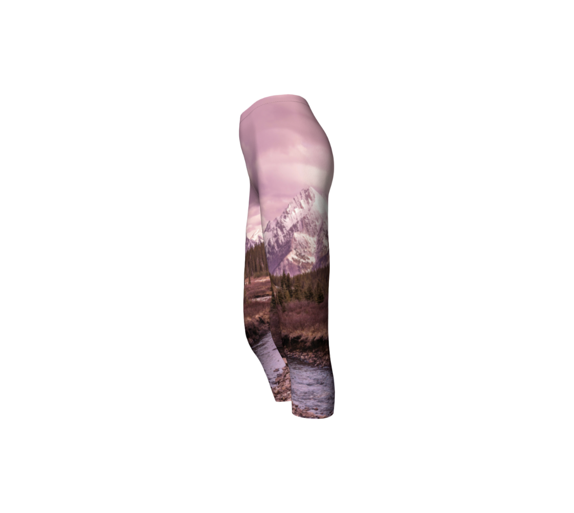 This is a side profile of a pair of 3/4 length capri leggings featuring a photo of a pink mountain scene. The image is of a snow covered mountain in the far distance with a meandering stream winding its way through the fall colored willow and forest of evergreen trees towards the mountain. A special filter used when taking this photograph created an over all pink hue to the image that really accentuated the sunset light at the time.