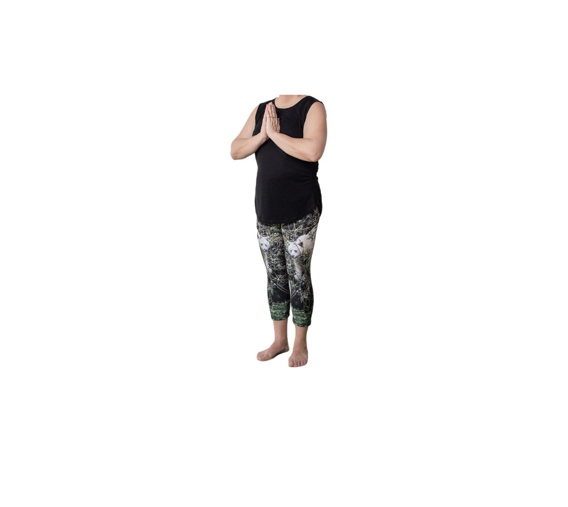 This is a front profile of a lady wearing a pair of 3/4 length capri leggings featuring a photo of a spirit bear. The background is green bushes and brown branches of the Great Bear Rainforest. The white spirit bear is stepping down out of the forest and is making her way to the stream below.