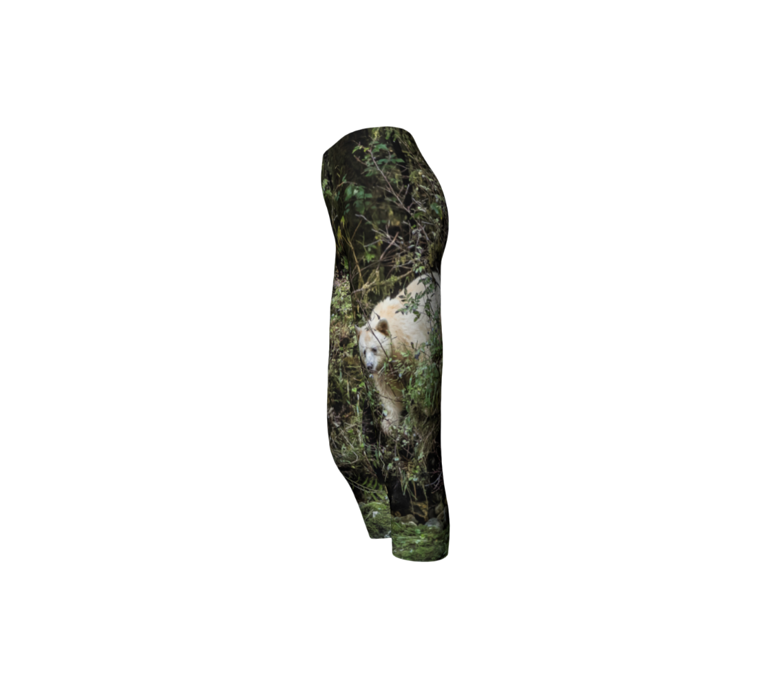 This is a side profile of a photo of a spirit bear on a pair of 3/4 length capri leggings. The background is green bushes and brown branches of the Great Bear Rainforest. The white spirit bear is stepping down out of the forest and is making her way to the stream below.