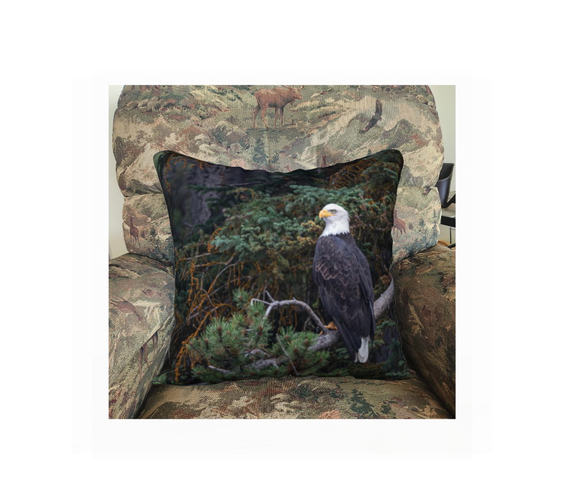 This 18 x 18 inch plush velveteen cushion cover features a photo of a mature bald eagle. The background is of trees and branches in various tones of green, gold, purple and grey. The bald eagle is perched on a fallen grey tree trunk with his back to us and its head is turned slightly, as he is looking towards you.  This image is of the cushion cover on a pillow form in a chair.