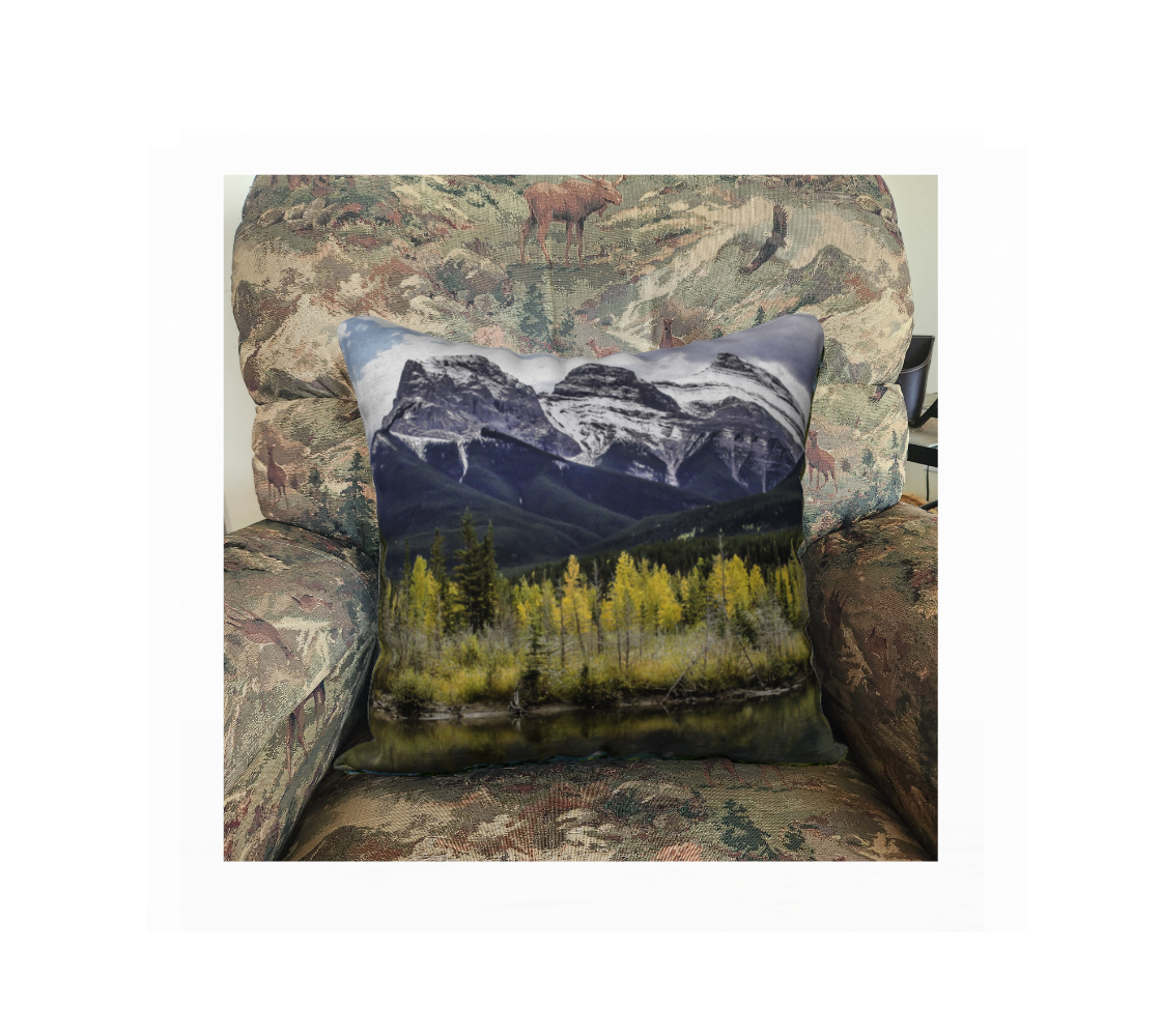This 18 x 18 inch plush velveteen cushion cover features a photo of an autumn scene of the Three Sisters Mountain with a little snow on the peaks. The cushion cover background is blue sky, clouds green tree on the mountain with yellow aspen trees in the foreground along the river's edge.. This image is of a cushion cover on a pillow form in a chair.