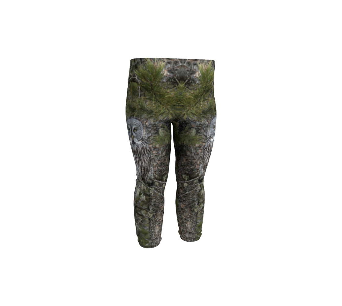 This is a front profile of infant leggings featuring a great grey owl on each side of the legs. The background is green pine boughs and lichen hanging from the grey pine tree branches, with a great grey owl with black, grey and white feathers, and yellow eyes, perched on a branch.