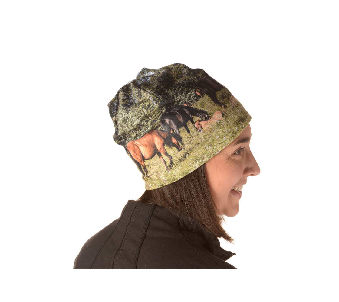 This is a side profile of a lady wearing a beanie featuring a photograph of wild foals lying in wildflowers in the summer, with a herd of wild horses grazing in a field surrounded by trees. Color tones include greens, browns, and black.